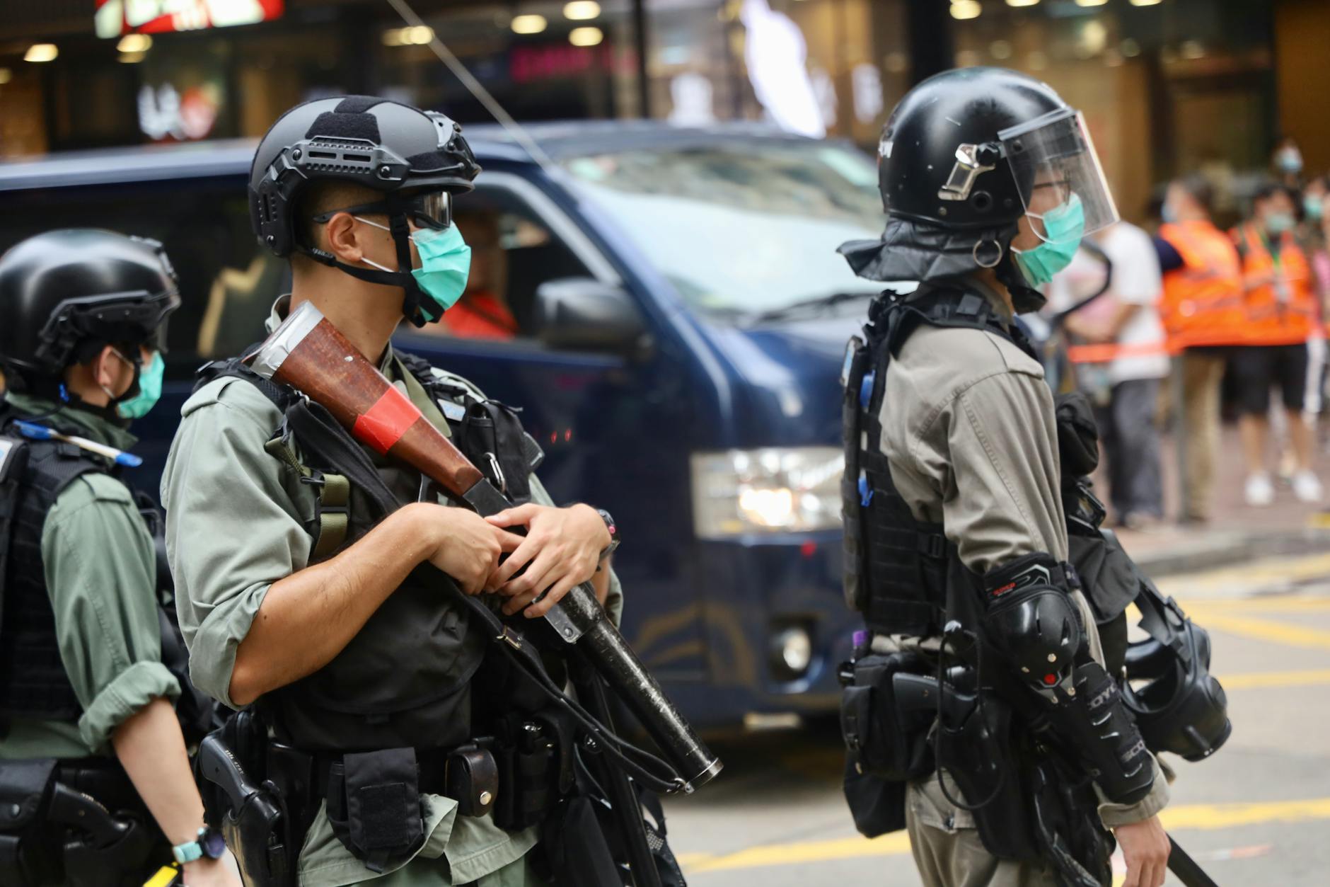 Police officers wearing riot gear and face masks stand in an urban street with protective equipment.