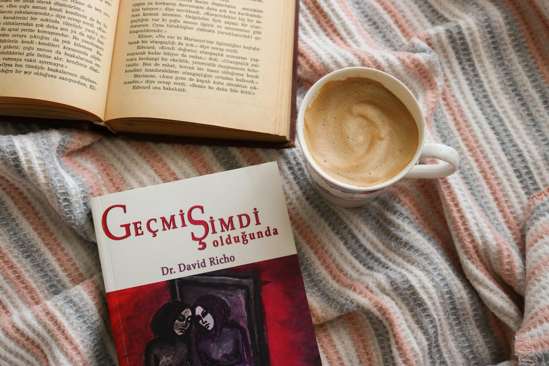 Flat lay of a cozy reading setup with books and a cup of coffee on a striped blanket.