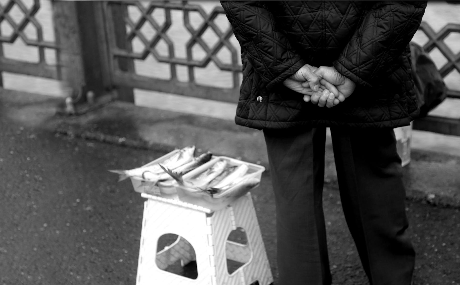 Monochrome scene of a street vendor with fish on a folding chair by the river.