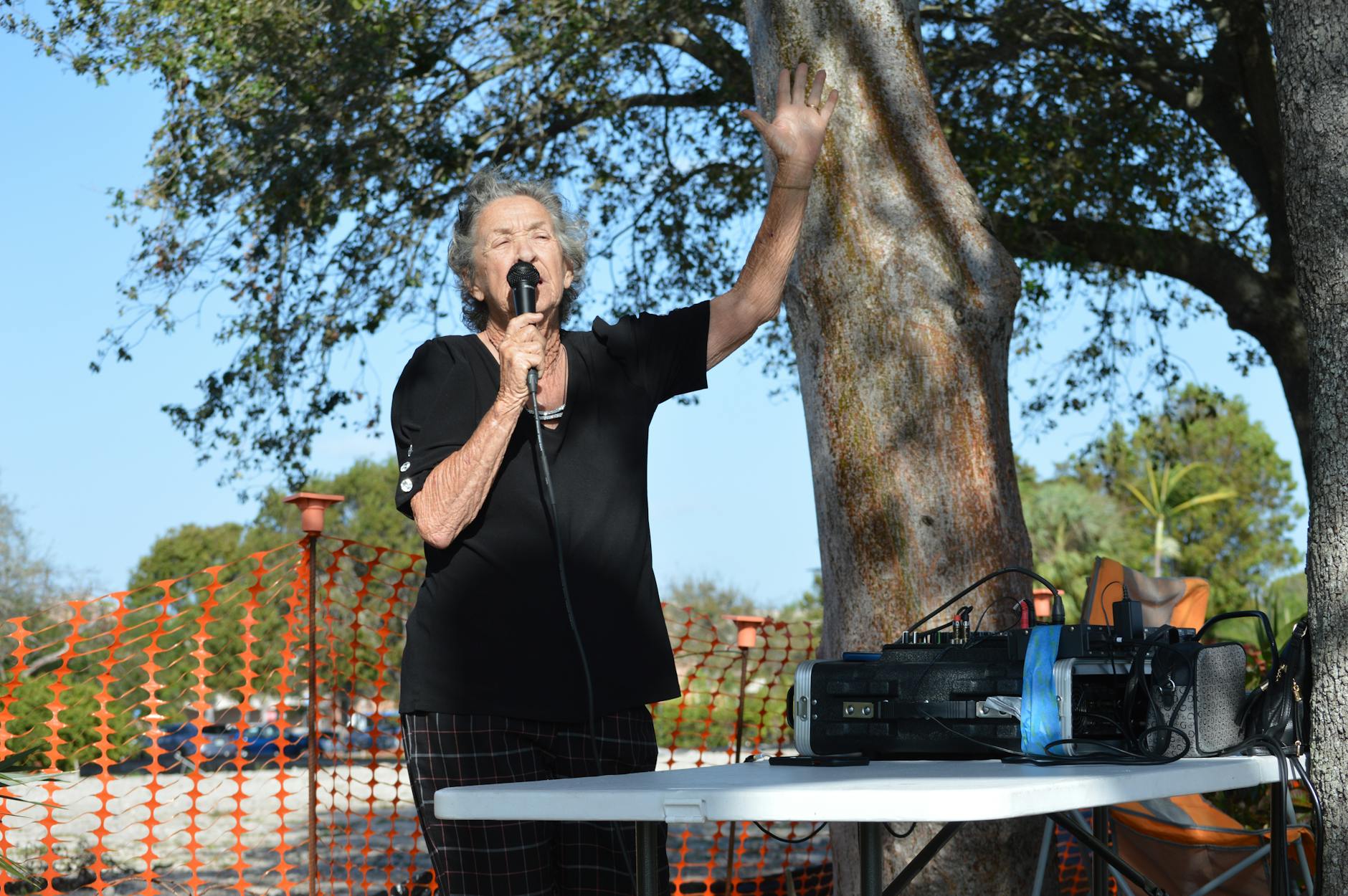 Elderly woman speaking into a microphone outdoors near sound equipment with a tree backdrop.