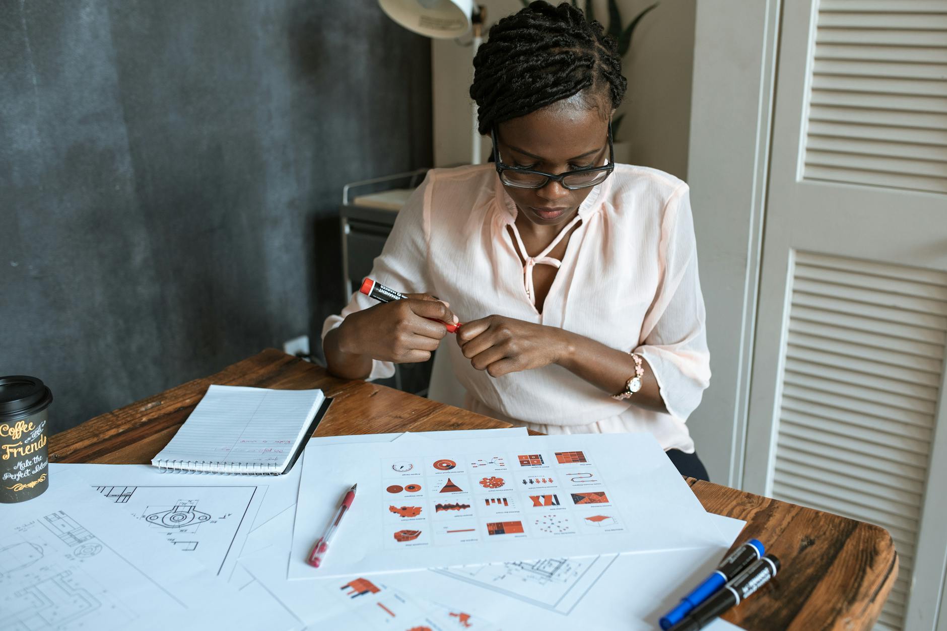 African woman with dreadlocks working on diagrams at an office desk, wearing eyeglasses and a blouse.
